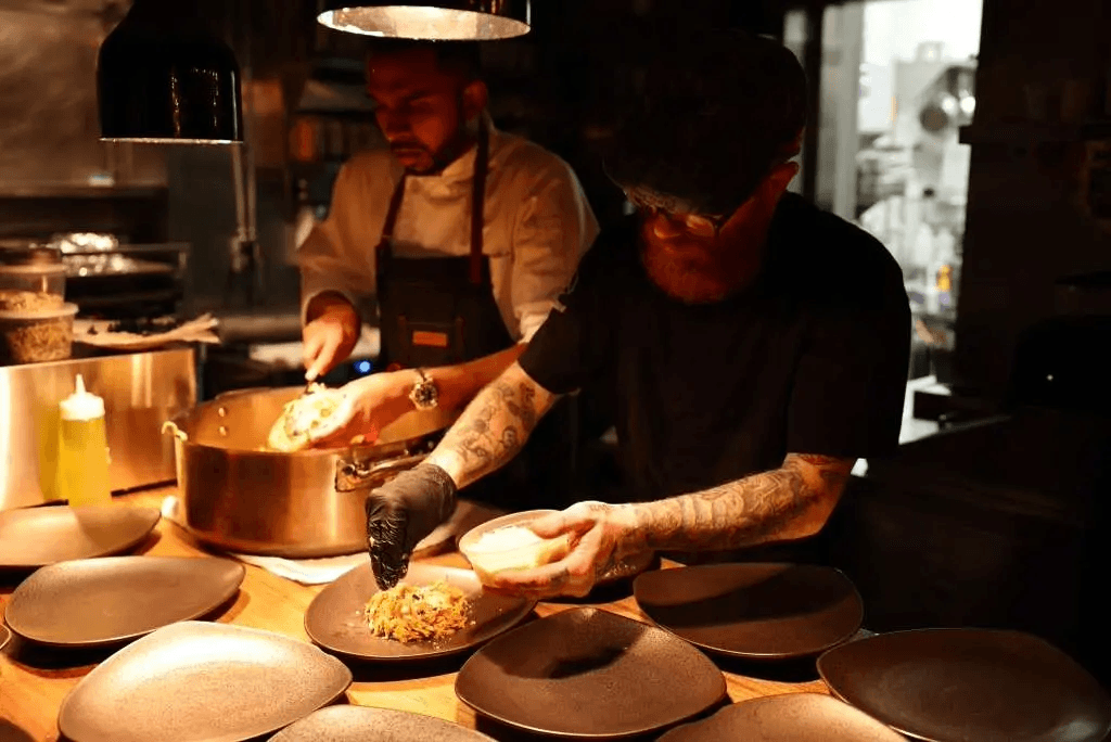 Two chefs plating dishes on a wood counter in a dimly lit professional kitchen, with warm overhead light.